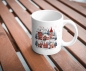 Preview: Weiße Keramiktasse mit einem charmanten Winterdorf-Motiv, elegant auf einem Holztisch präsentiert, ideal für gemütliche Momente während der Weihnachtszeit.
